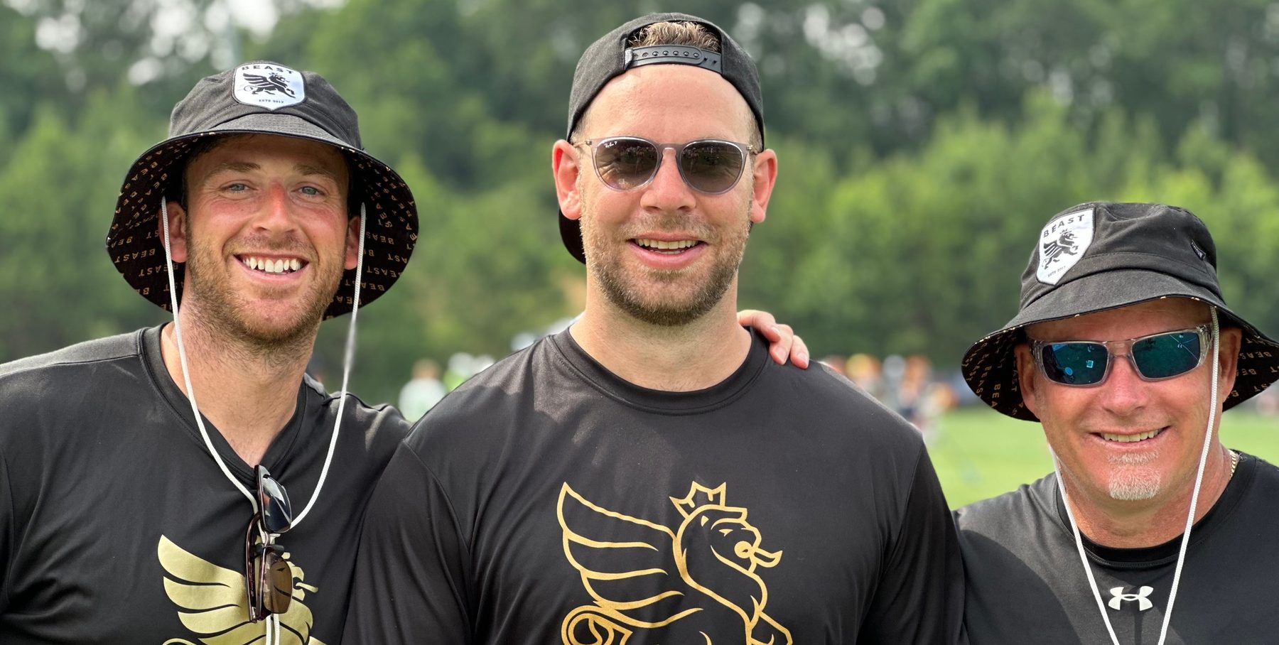 Three BEAST coaches in black BEAST shirts and bucket hats smiling on the sideline at a tournament